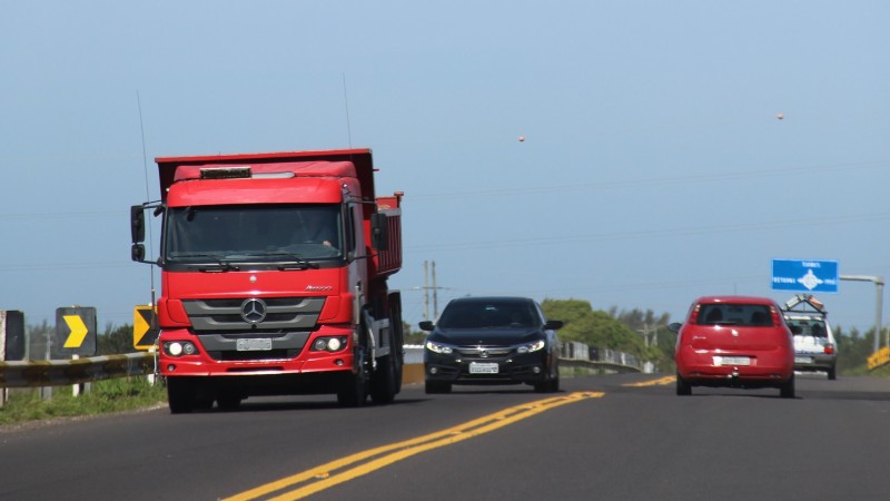 A foto mostra um caminhão e diversos carros trafegando nos dois sentidos de uma rodovia pavimentada.