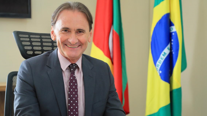 A foto mostra o secretário de Logística e Transportes do Estado, Juvir Costella, sentado à mesa, com bandeiras do Brasil e do Rio Grande do Sul ao fundo.