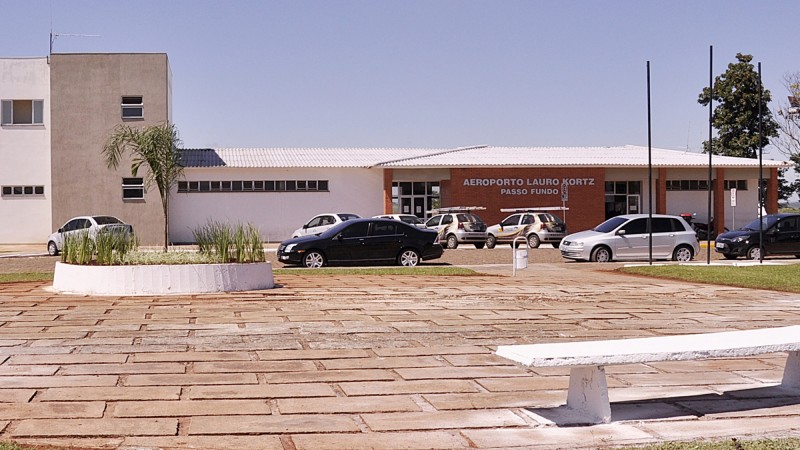 A foto mostra a fachada do Aeroporto Lauro Kortz, de Passo Fundo, com carros estacionados na frente.