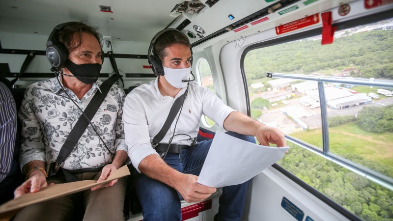A foto mostra o governador Eduardo Leite e o secretário Juvir Costella dentro de um helicóptero, olhando projetos, enquanto sobrevoam a ERS-118, na Região Metropolitana de Porto Alegre