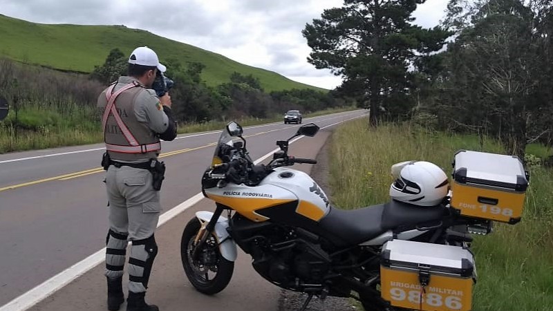 A foto mostra um policial rodoviário operando um radar portátil, no acostamento de uma estrada, com uma moto estacionada ao lado. Ao longe, um carro passa pela rodovia.