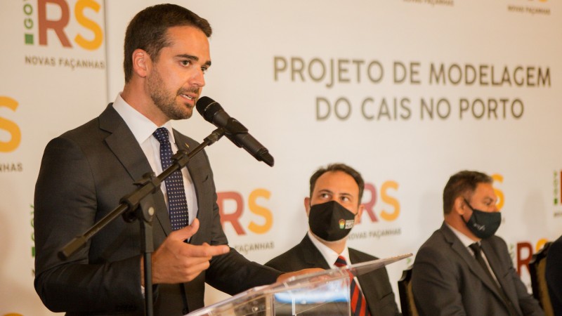 A foto mostra o governador Eduardo Leite discursando ao microfone, com uma tapadeira atrás com os escritos "Projeto de Modelagem do Cais do Porto"