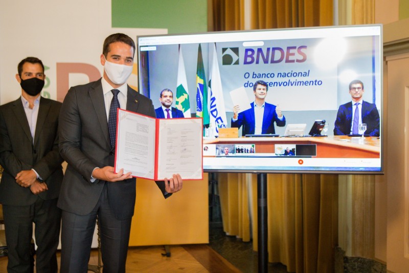 A foto mostra o governador Eduardo Leite mostrando um documento oficial assinado, tendo ao fundo uma tela de videochamada em que aparece o presidente do BNDES.
