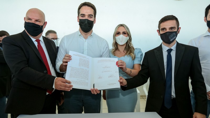 A foto mostra o governador do Estado, Eduardo Leite, mostrando o documento oficial de assinatura de convênio para pavimentação do Caminho do Pão e do Vinho, junto com prefeitos da região.