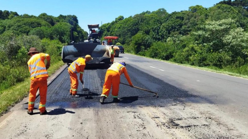 Três trabalhadores colocam asfalto em pista com árvores nas laterais, tendo ao fundo máquinas e veículos de transporte.
