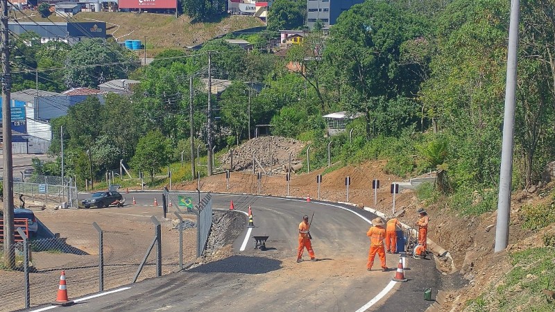 Trabalhadores em uniformes laranja trabalham na marcação com tinta em pista de asfalto em curva para a esquerda com árvores e casas nas laterais. 