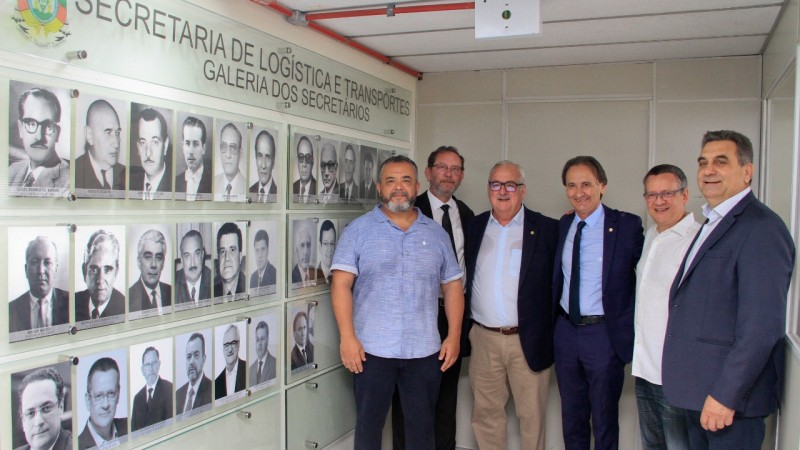 Cinco ex-secretários de Transporte e o atual secretário posam ao lado de mural de fotografias em parede.