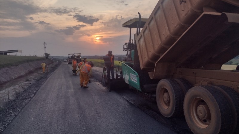 Caminhão despeja asfalto na pista, e trabalhadores espalham material. Ao fundo, sol aparece entre nuvens.