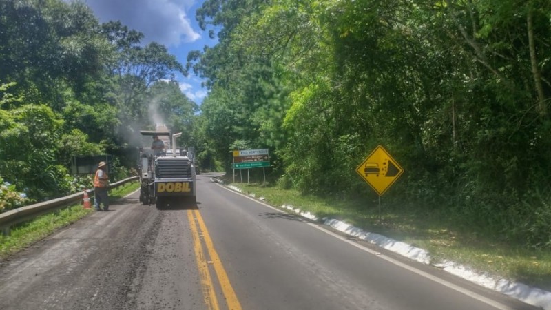 Imagem mostra trabalhadores operando máquinas em estrada em obras, com vegetação nas laterais e placa à direita sinalizando possibilidade de desmoronamento.