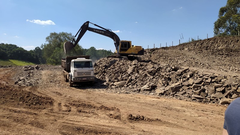 Máquina coloca pedras em caminhão estacionado em pista de terra.