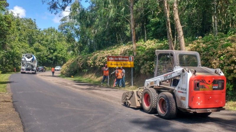 Trabalhadores operam máquinas preparando pista para receber manutenção asfáltica. Árvores nas laterais da pista.