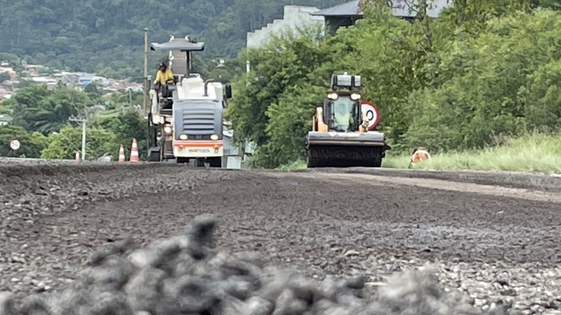 Máquinas trabalham em pista que recebe asfalto. Árvores e vegetação nas laterais da pista e ao fundo da imagem.