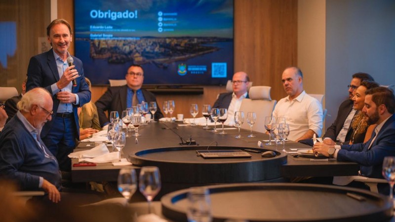 Na foto, o secretário Costella é um homem branco, de cabelos escuros, fala ao microfone em um evento, observado pelos demais presentes.