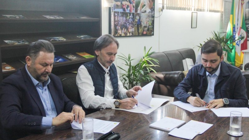 Três homens brancos assinam documento sobre uma mesa com papeis e copos com água. Os homens são o vice-governador do Rio Grande do Sul Gabriel Souza, o secretário de Logística e Transportes do estado, Juvir Costella e o prefeito de Estrela, Elmar Schneider.