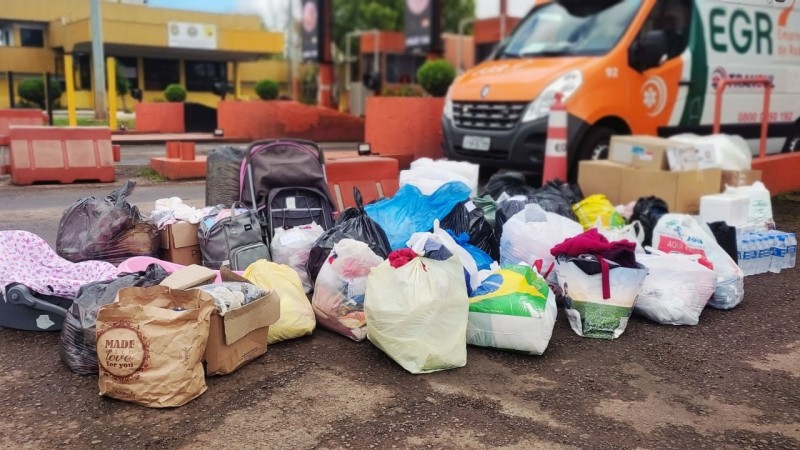Imagem mostra sacos com itens doados para vítimas das enchentes em praça de pedágio, com ônibus da EGR ao fundo