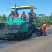 Trabalhadores operam máquinas para colocação de asfalto em pista com árvores ao fundo.