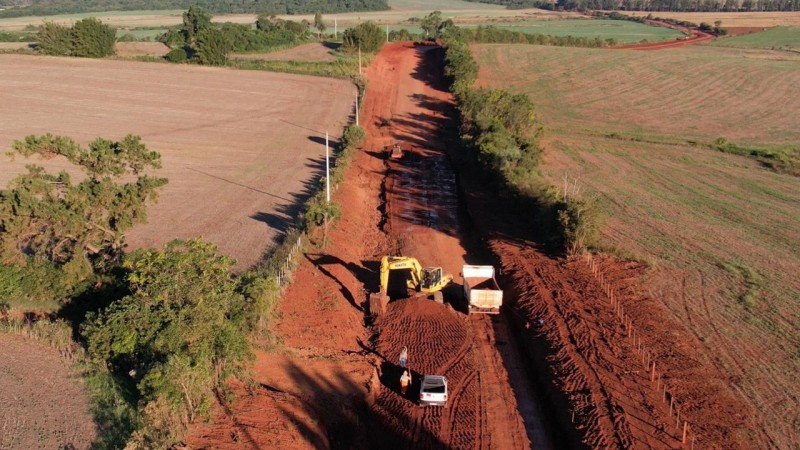 Obra de pavimentação em uma estrada de terra. Na imagem, aparecem uma retroescavadeira, um caminhão, um automóvel e trabalhadores.