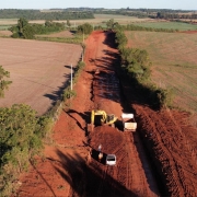 Obra de pavimentação em uma estrada de terra. Na imagem, aparecem uma retroescavadeira, um caminhão, um automóvel e trabalhadores. 